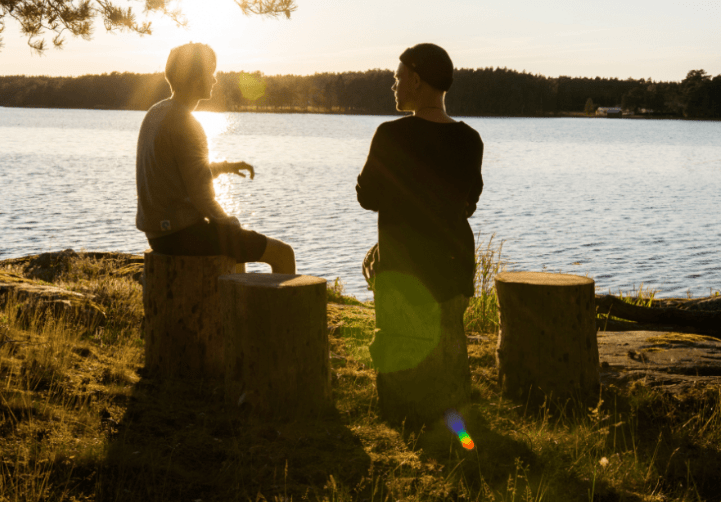 Zwei Menschen, die an einem See sitzen und sich unterhalten, während die Sonne untergeht. Es ist wichtig, dass Sie Ihren Mitarbeitern und Ihrem Team eine Stimme geben, damit eine Grundlage für ein gesundes Arbeitsumfeld geschaffen werden kann. Mentoring ist ein mögliches Konzept dafür.